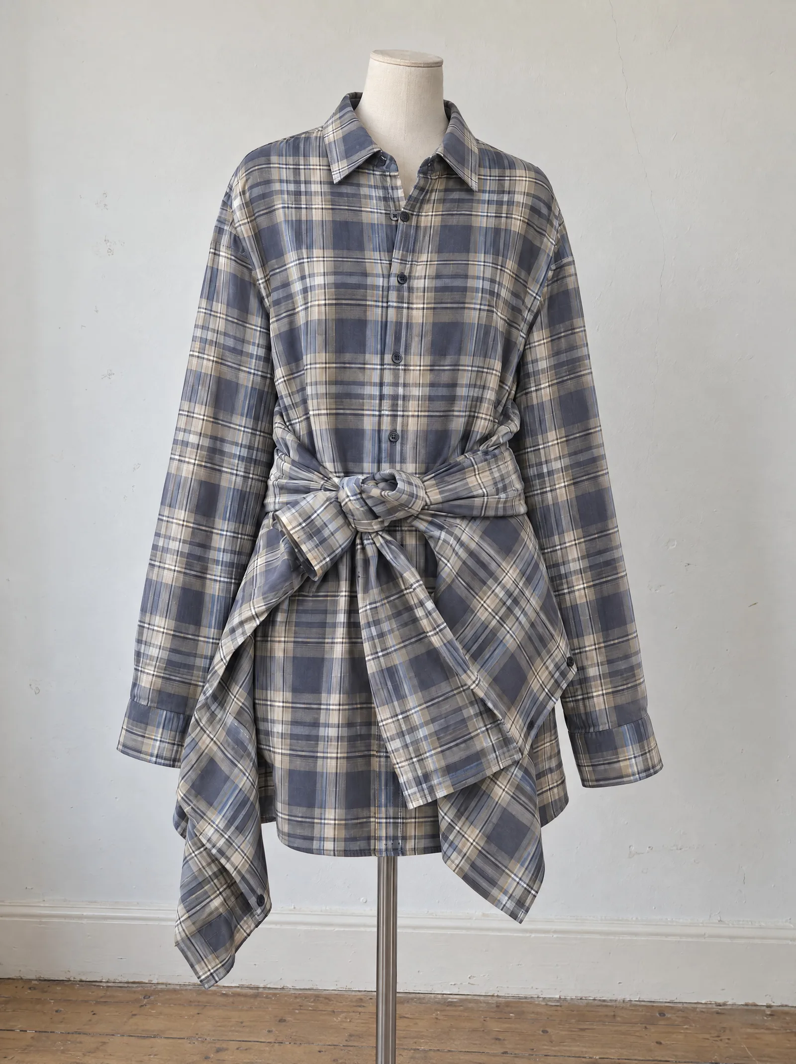 The plaid shirt dress on a dressmaker's bust standing slightly off centre in an ordinary room, against a plain painted wall with faint scuffs and a hairline crack